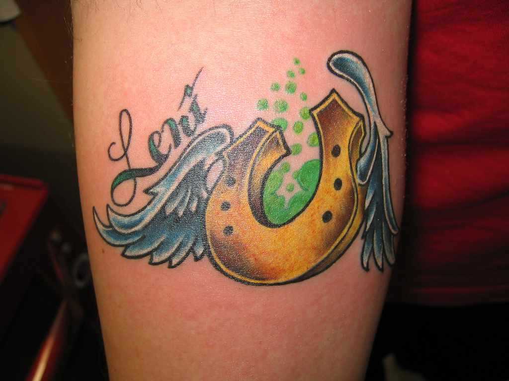 Tatuagem no ombro da menina - ferradura com asas e inscrição
